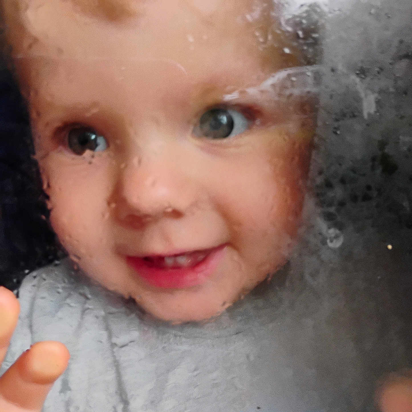 Mattie is registered to the contest to win money with this photo: child, toddler, portrait, face, eyes, smiling, window, raindrops, glass, outdoor, close_up, blond_hair, blue_eyes, hand, gray_clothing, baby, curious, looking_at_camera, natural_light, photography