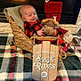 baby, plush_toy, basket, fireplace, christmas_stocking, plaid_pajamas, blanket, wooden_sign, holiday_decor, indoor, smiling, child, cute, festive, toy, cozy, warm, sleigh, red, black