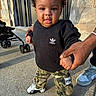 adidas, adult_hands, baby, black_sweatshirt, camo_pants, casual_clothing, child, curly_hair, cute, holding_hands, outdoor, person, portrait, sidewalk, sneakers, standing, stroller, sunlight, toddler, urban