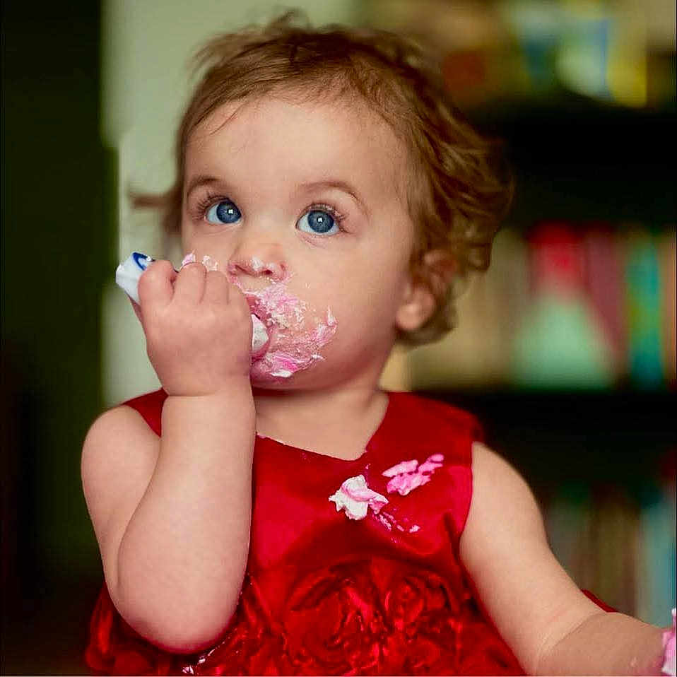 Ember is registered to the contest to win money with this photo: baby, background_blur, blue_eyes, bookshelf, child, close_up, curly_hair, cute, eating, face, food, hand, happy, indoor, messy_face, person, pink_frosting, portrait, red_dress, toddler