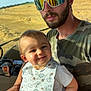 baby, man, child, bib, sunglasses, beard, camouflage_shirt, vehicle_interior, field, sunlight, window, smile, holding, portrait, person, nature, daylight, happy, casual_clothing, dinosaur_print