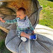 Michel a rejoint le concours — aidez-le/la à gagner de superbes lots ! toddler, child, pacifier, bib, plaid_shirt, plaid_shorts, blue_shoes, wooden_sculpture, flower_shape, outdoor, grass, sunlight, lens_flare, reaching_hand, nature, daylight, smiling, cute, sitting, playful