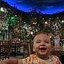 Milo is registered to the contest to win money with this photo: baby, child, smiling, happy, high_chair, pumpkin_shirt, halloween, indoor, restaurant, wooden_chairs, table, decor, plants, greenery, starry_ceiling, night, portrait, person, infant, cute