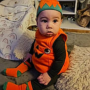 Bailey is registered to the contest to win money with this photo: baby, child, pumpkin_costume, orange, black_clothing, socks, hat, indoor, living_room, blanket, pillow, carpet, furniture, couch, floor, person, cute, seated, portrait, costume