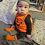 Bailey is registered to the contest to win money with this photo: baby, child, pumpkin_costume, orange, black_clothing, socks, hat, blanket, pillow, sofa, living_room, carpet, cushion, furniture, indoors, person, cute, expression, sitting, floor