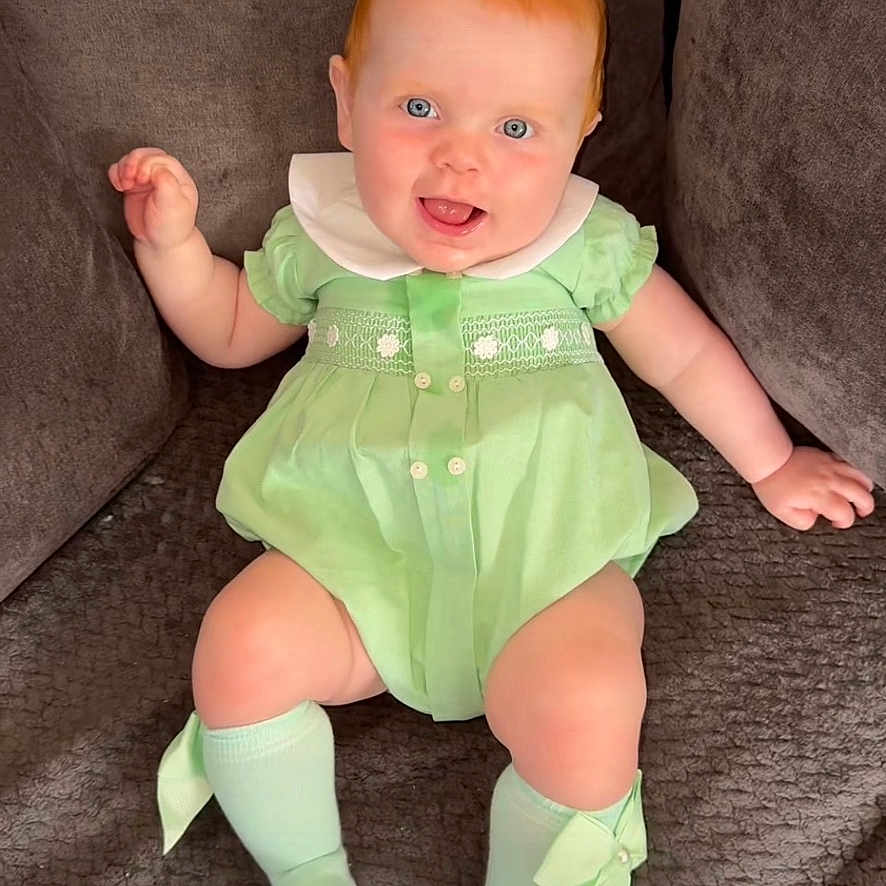 Isla-Rose joined the competition — help win amazing prizes! apparel, baby, blue_eyes, bow, child, comfortable, couch, cute, fabric, green_dress, happy, indoor, infant, person, plush, red_hair, sitting, smiling, socks, tongue_out