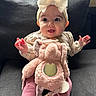 baby, child, bow, headband, plush_toy, elephant_toy, couch, pink_clothing, smiling, happy, indoors, cute, portrait, sitting, soft_toy, infant, hands, face, cozy, warm_lighting
