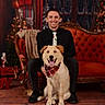 man, dog, smiling, red_couch, plaid_bandana, holiday_decor, vintage_furniture, indoor, portrait, festive, black_clothing, wood_floor, window, smiling_dog, happy, seated, tie, warm_lighting, decorations, cheerful