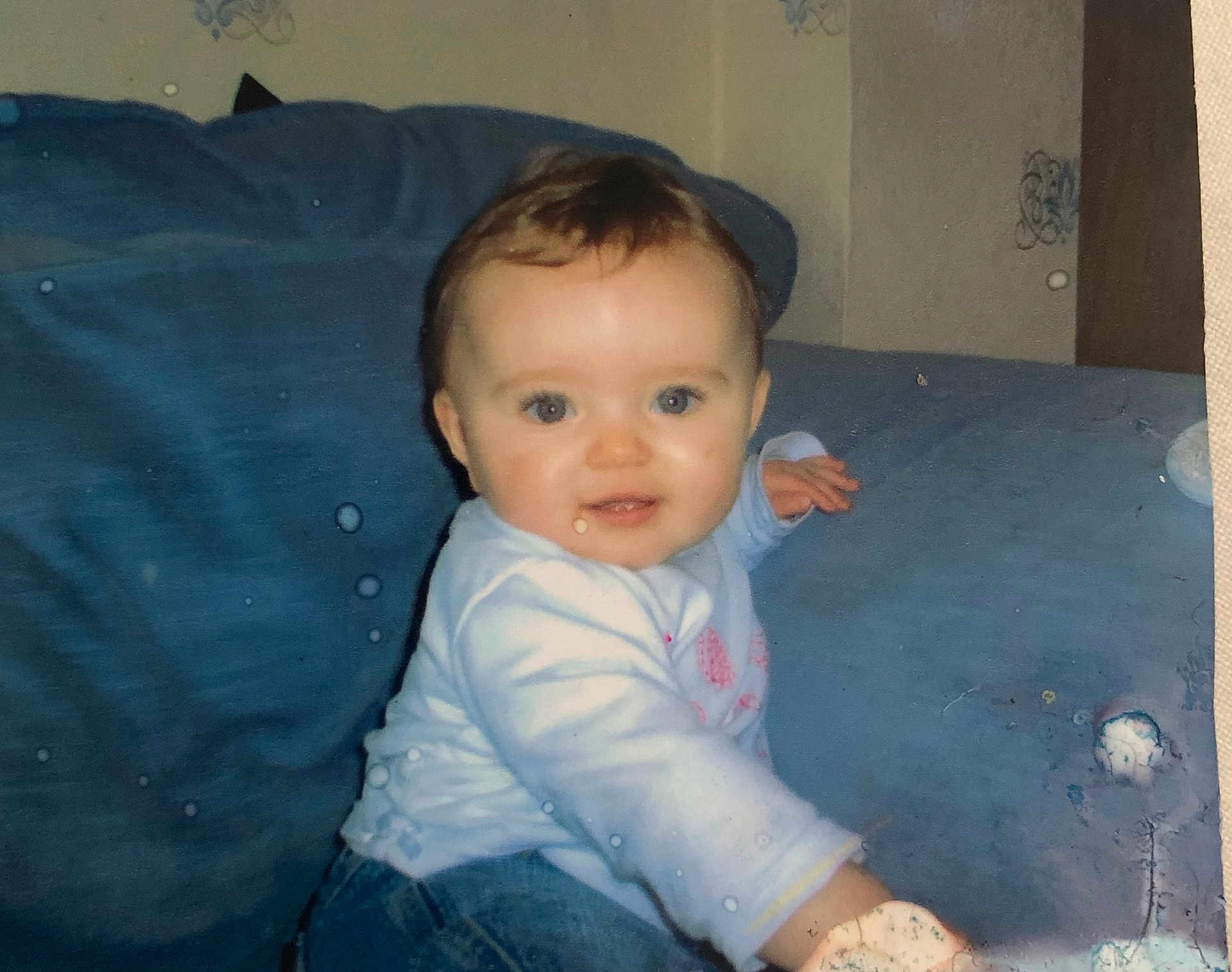 Taylor is registered to the contest to win money with this photo: baby, child, blue_couch, indoor, smile, face, person, clothing, jeans, long_sleeve, young_child, cute, sitting, home, furniture, portrait, infant, happy, reaching, photograph