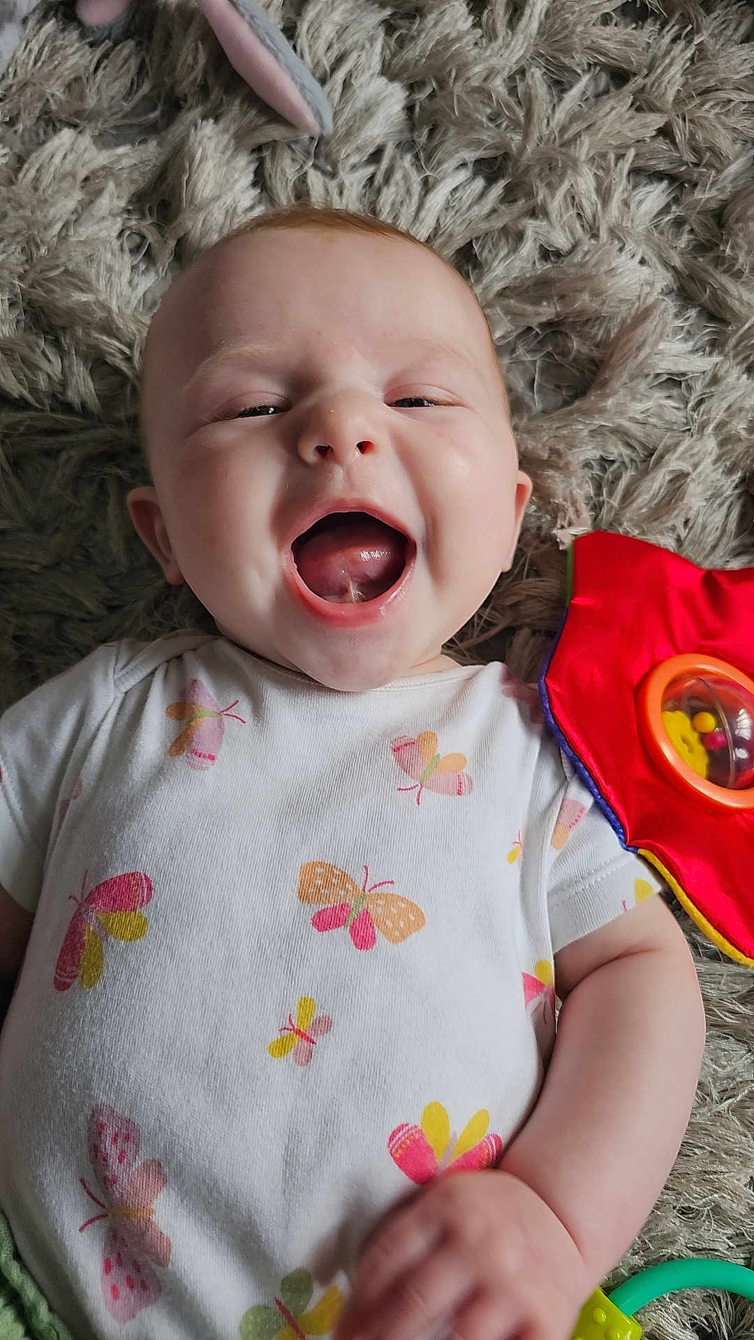 Ruby joined the competition — help win amazing prizes! baby, smiling, infant, onesie, butterfly_pattern, toy, red, green, yellow, floor_rug, happy, cute, child, playtime, closeup, skin, hand, mouth_open, face, lying_down