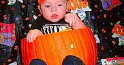 Orion is registered to the contest to win money with this photo: baby, infant, child, pumpkin, halloween, orange_hat, black_clothing, sitting, portrait, face, hands, curious, surprised, colorful, patterned_blanket, spooky, cartoon, indoor, date_stamp, seasonal