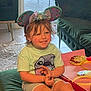 child, smile, mouse_ears, headband, tshirt, shorts, indoor, cushion, carpet, furniture, sofa, table, playful, happy, casual_clothing, home, person, cute, young, relaxed