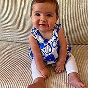 Laura a rejoint le concours — aidez-le/la à gagner de superbes lots ! baby, child, smiling, sitting, couch, clothing, white_leggings, blue_flower_top, indoors, happy, face, skin, person, cute, infant, foot, hand, furniture, portrait, young