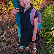 Mia is registered to the contest to win money with this photo: child, smiling, outdoor, backpack, grass, plants, post, happy, girl, sunlight, shoes, dress, nature, portrait, person, feet, greenery, casual, standing, daylight