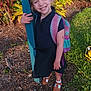 child, smiling, outdoor, backpack, grass, plants, post, happy, girl, sunlight, shoes, dress, nature, portrait, person, feet, greenery, casual, standing, daylight