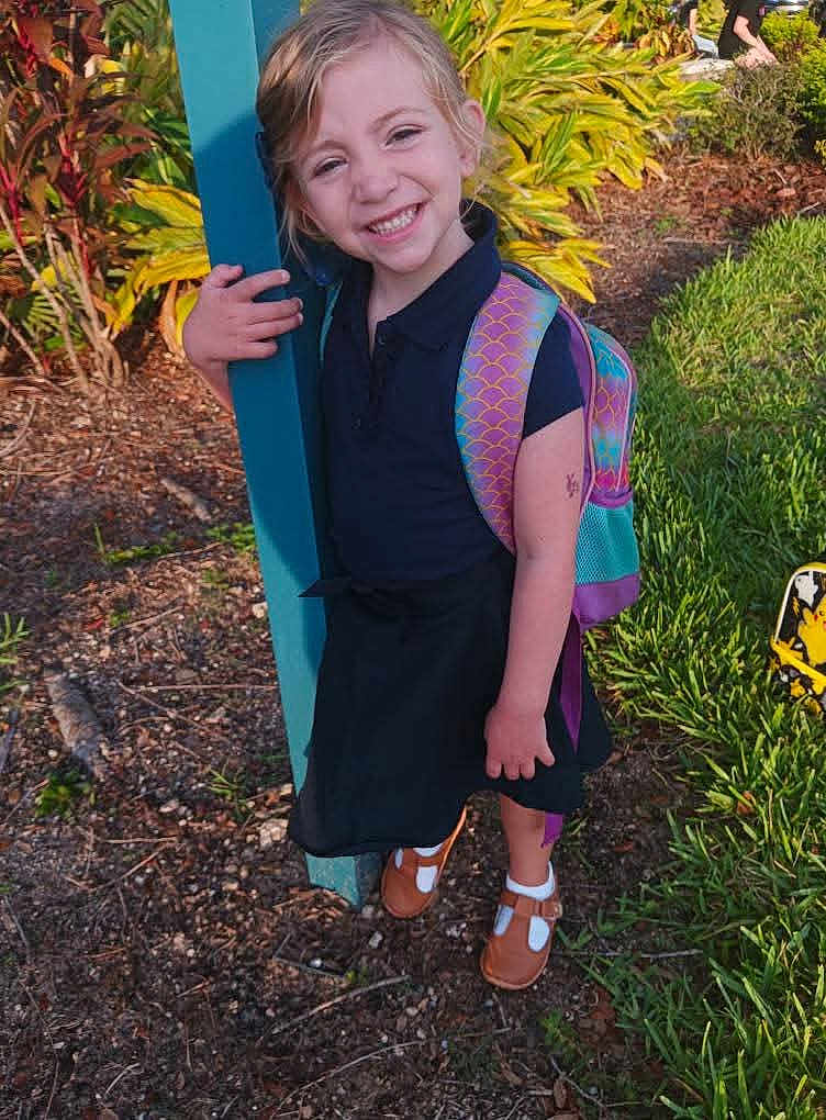 Mia is registered to the contest to win money with this photo: child, smiling, outdoor, backpack, grass, plants, post, happy, girl, sunlight, shoes, dress, nature, portrait, person, feet, greenery, casual, standing, daylight