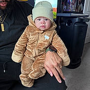 Gianni joined the competition — help win amazing prizes! baby, child, adult, beanie, brown_clothing, fuzzy_suit, indoor, person, hand, watch, facial_expression, computer_screen, text, gaming, chair, carpet, room, decor, portrait, casual