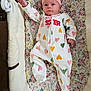 baby, infant, pink_bow, onesie, hearts, floral_pattern, cushion, lying_down, face, hand, footed_pajamas, text, sleepwear, child, cute, indoor, portrait, soft_toys, newborn, headband