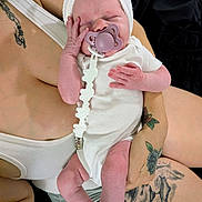 Line is registered to the contest to win money with this photo: baby, newborn, pacifier, white_onesie, headband, sleeping, tattoo, arm, hand, skin, person, infant, cute, peaceful, indoor, closeup, holding, human, adult, tender