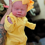 Line is registered to the contest to win money with this photo: baby, infant, yellow_outfit, headband, bow, tattoo, hand, pacifier_clip, squinting, expression, indoor, person, closeup, cute, newborn, clothing, holding, soft_light, portrait, human