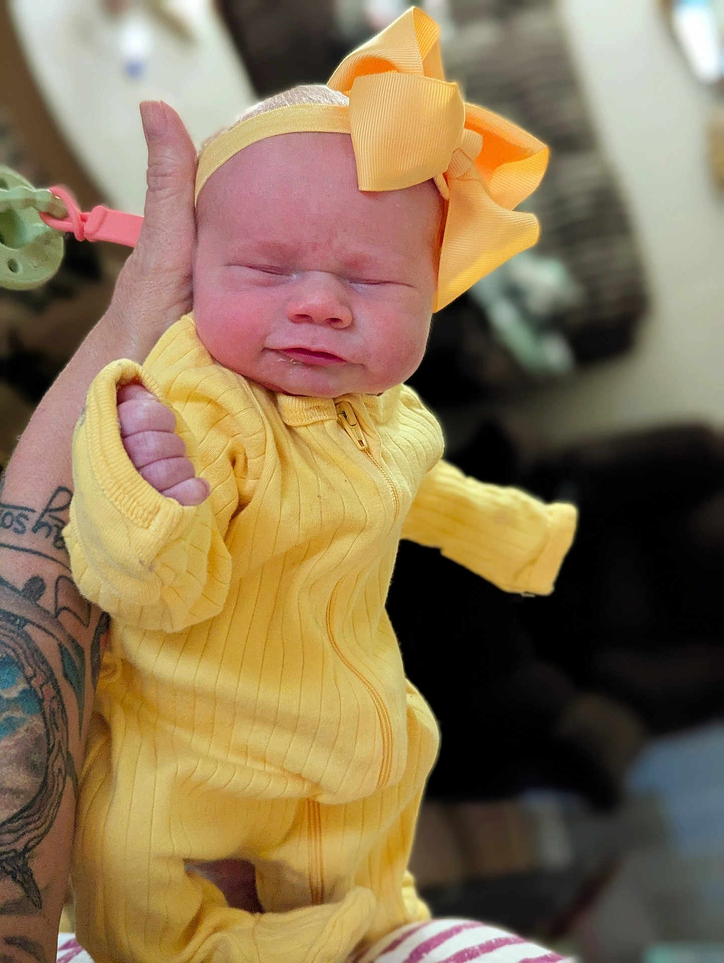 Line is registered to the contest to win money with this photo: baby, infant, yellow_outfit, headband, bow, tattoo, hand, pacifier_clip, squinting, expression, indoor, person, closeup, cute, newborn, clothing, holding, soft_light, portrait, human