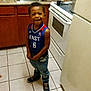 boy, cabinet, can, casual_clothing, child, countertop, cup, happy, indoor, jeans, kitchen, person, plastic_bottle, sink, smiling, sneakers, stove, tile_floor, water_bottle, young