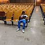 auditorium, blue_pants, boy, brick_wall, casual, child, concrete_floor, empty_seats, indoor, large_room, lighting, quiet, relaxed, seated, single_person, sneakers, spacious, white_shirt, wooden_chairs, young_person