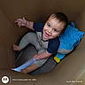 baby, child, smiling, happy, portrait, indoor, cardboard_box, sitting, lying_down, waving, blue_pillow, toy, colorful, casual_clothing, short_hair, barefoot, candid, photography, warm_lighting, playful