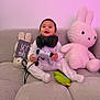 Noham a rejoint le concours — aidez-le/la à gagner de superbes lots ! baby, child, headphones, game_controller, plush_toy, bunny, couch, gray_couch, pink_light, smiling, sitting, indoors, cozy, soft_toy, cute, person, infant, pajamas, happy, toy