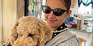 Phoenix is registered to the contest to win money with this photo: person, dog, indoor, kitchen, sunglasses, curly_hair, hoodie, pearl_necklace, wooden_cabinets, refrigerator, stool, counter, smile, pet, holding, casual_clothing, curtains, cup, tableware, flooring