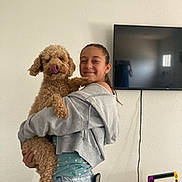 Phoenix is registered to the contest to win money with this photo: girl, dog, indoor, hugging, smiling, curly_fur, television, gaming_console, casual_clothing, jeans, hoodie, wooden_stool, remote_control, happy, pet, playful, home, technology, living_room, cord