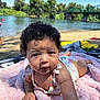 baby, child, curly_hair, blanket, beach, river, bridge, trees, water, outdoor, sunlight, blue_sky, sand, person, infant, reaching, portrait, nature, summer, cute