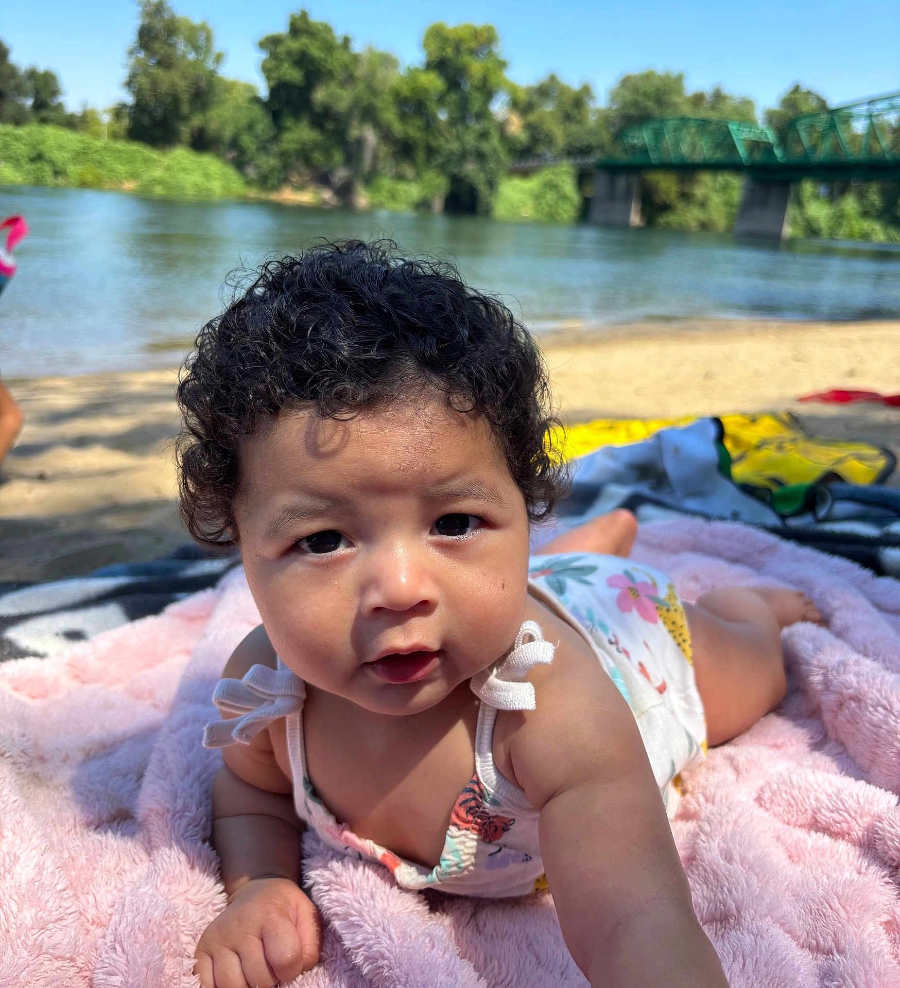 Zaiyla joined the competition — help win amazing prizes! baby, child, curly_hair, blanket, beach, river, bridge, trees, water, outdoor, sunlight, blue_sky, sand, person, infant, reaching, portrait, nature, summer, cute