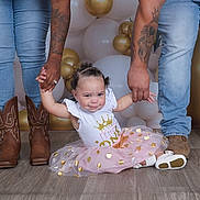 Alayna is registered to the contest to win money with this photo: toddler, child, baby, jeans, boots, tattoos, balloons, pink_tutu, floor, hands, adults, celebration, party, family, holding_hands, curly_hair, white_shirt, gold, indoor, smile