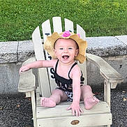Stella is registered to the contest to win money with this photo: baby, child, outdoor, grass, chair, wooden_chair, hat, sunhat, smiling, happy, sitting, swimsuit, leopard_print, concrete, stone_wall, summer, cute, infant, person, playful