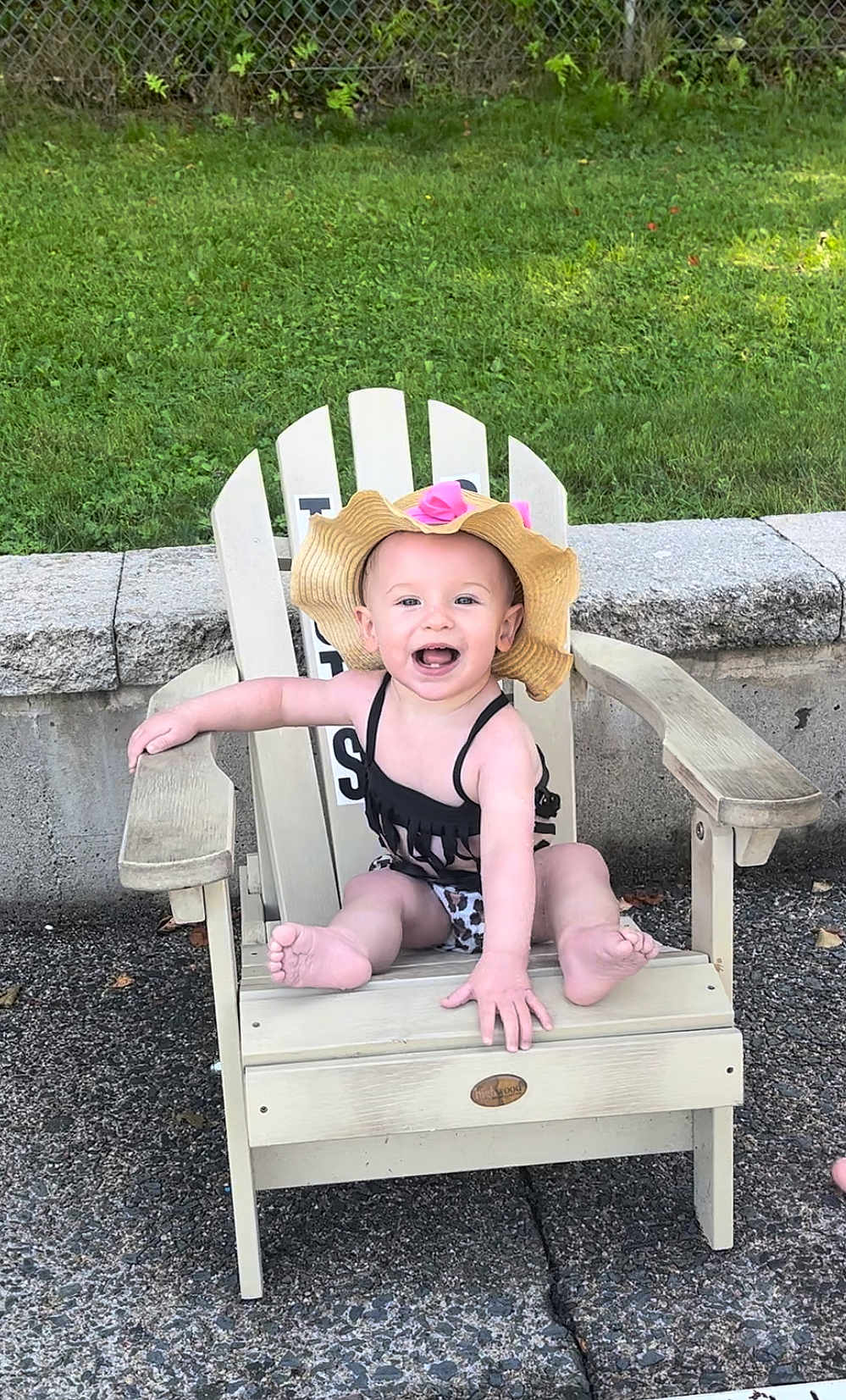 Stella is registered to the contest to win money with this photo: baby, child, outdoor, grass, chair, wooden_chair, hat, sunhat, smiling, happy, sitting, swimsuit, leopard_print, concrete, stone_wall, summer, cute, infant, person, playful