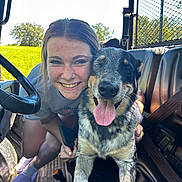 Toby is registered to the contest to win money with this photo: woman, dog, smiling, tongue_out, vehicle_interior, seat, seatbelt, steering_wheel, grass, trees, sunny, daylight, casual_clothing, shorts, socks, slippers, happy, pet, friendship, outdoor
