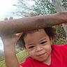 child, curly_hair, red_shirt, outdoor, metal_bars, grass, wooden_post, face, person, nature, playful, young_child, expression, curious, mischievous, daylight, sky, tree, hand, portrait
