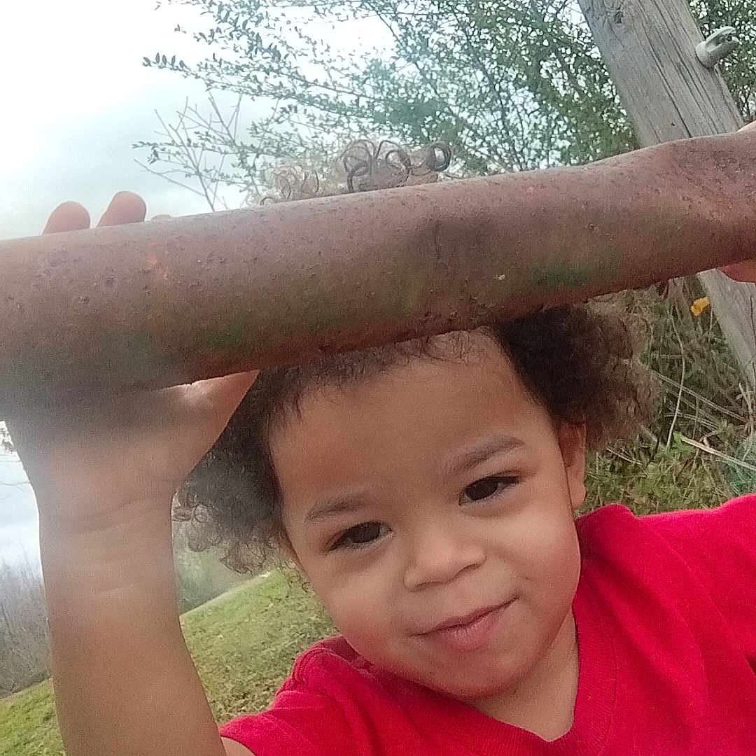 Christian joined the competition — help win amazing prizes! child, curly_hair, red_shirt, outdoor, metal_bars, grass, wooden_post, face, person, nature, playful, young_child, expression, curious, mischievous, daylight, sky, tree, hand, portrait