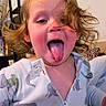 child, toddler, tongue_out, dinosaur_onesie, cute, playful, indoor, hair, face, expression, funny, home, furniture, bookshelf, person, portrait, smiling, clothing, young_child, happy