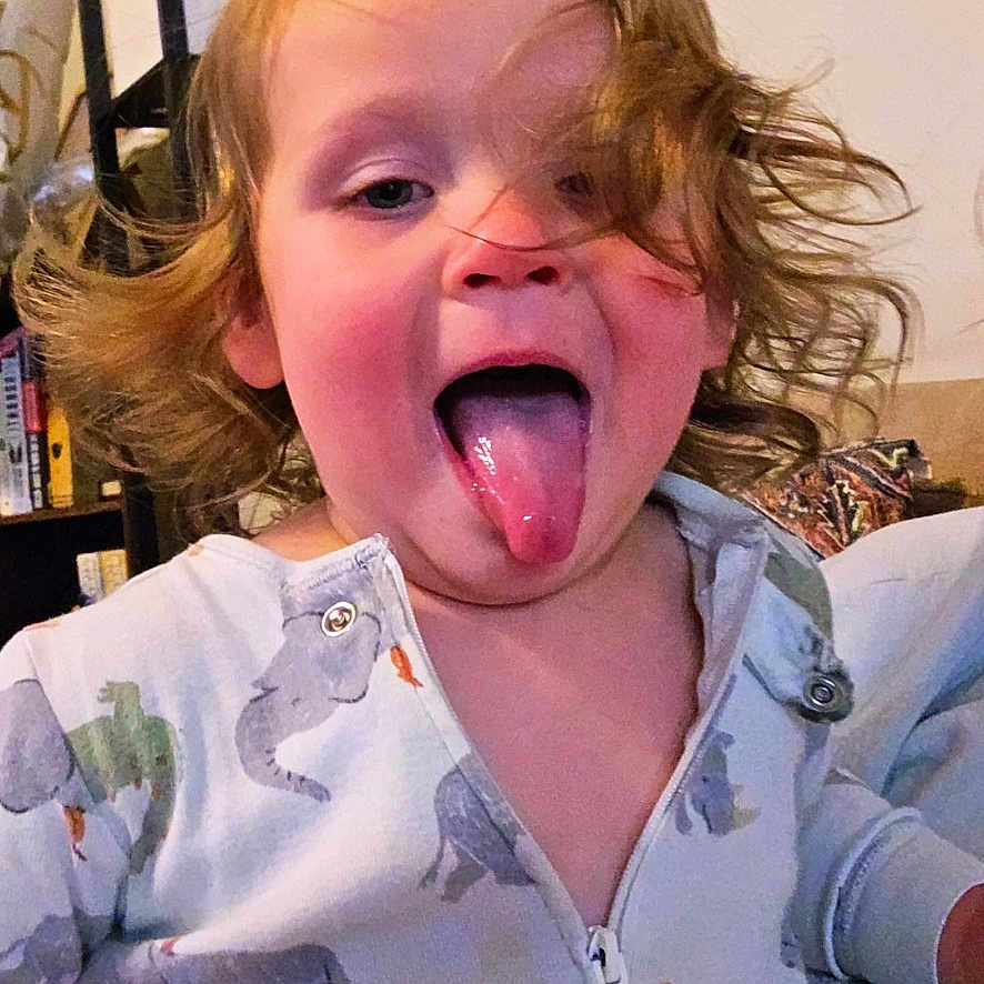 Connor joined the competition — help win amazing prizes! child, toddler, tongue_out, dinosaur_onesie, cute, playful, indoor, hair, face, expression, funny, home, furniture, bookshelf, person, portrait, smiling, clothing, young_child, happy