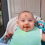 Ryker joined the competition — help win amazing prizes! baby, high_chair, bib, messy_face, food, orange_sauce, smiling, indoor, toy, ambulance_print, door, child, happy, cute, infant, feeding_time, playful, face, clothing, person