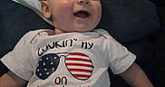 Ryker joined the competition — help win amazing prizes! baby, blanket, blue, celebration, child, clothing, crib, face, flag, fourth_of_july, happy, holiday, infant, lying_down, onesie, portrait, red, smiling, stars, white