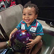 Ramsey is registered to the contest to win money with this photo: toddler, child, smiling, bowling_ball, indoor, chair, curly_hair, colorful_shirt, hands, adult, happy, sports, recreation, leisure, person, seat, ball, fun, playing, family