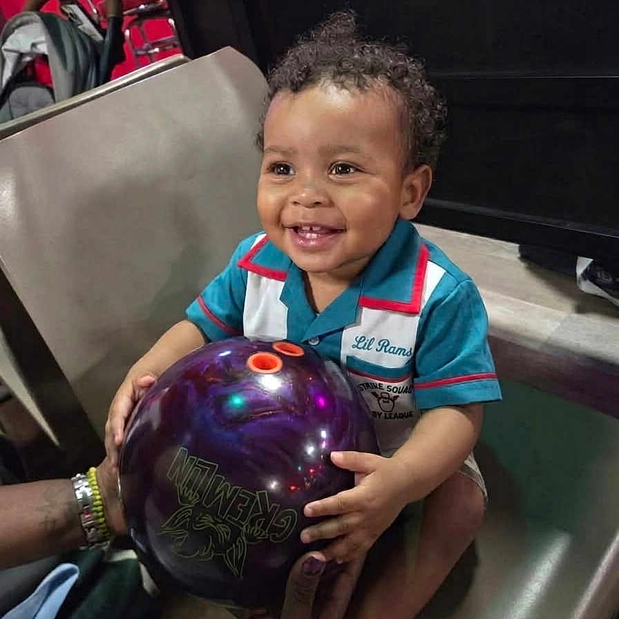 Ramsey is registered to the contest to win money with this photo: toddler, child, smiling, bowling_ball, indoor, chair, curly_hair, colorful_shirt, hands, adult, happy, sports, recreation, leisure, person, seat, ball, fun, playing, family