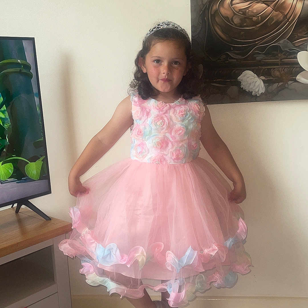 Isabella joined the competition — help win amazing prizes! buddha, child, curly_hair, dress, floor, flooring, furniture, girl, indoor, painting, pastel_colors, pink_dress, portrait, pose, sandals, smiling, television, tiara, wall, wooden_table