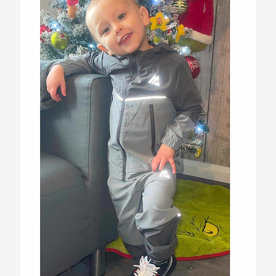 Harry is registered to the contest to win money with this photo: child, christmas_tree, couch, decorations, festive, gray_clothing, grinch, happy, holiday_decor, holiday_season, home, indoor, lights, ornaments, pose, reflective_clothing, smiling, sneakers, wood_floor, young_child