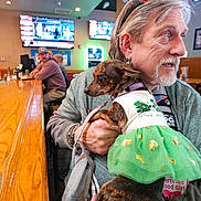 Kona Marie is registered to the contest to win money with this photo: man, dog, bar, indoor, clothing, green_dress, pet, person, holding, earrings, television, beard, table, seated, hat, casual, leash, furniture, light, social