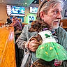 man, dog, bar, indoor, clothing, green_dress, pet, person, holding, earrings, television, beard, table, seated, hat, casual, leash, furniture, light, social