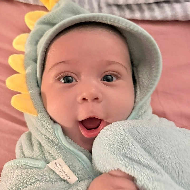 Enzo is registered to the contest to win money with this photo: baby, child, hooded_towel, dinosaur_costume, surprised, cute, soft_texture, blue_towel, yellow_spikes, indoor, portrait, infant, clothing, face, expression, hand, bed, blanket, happy, closeup